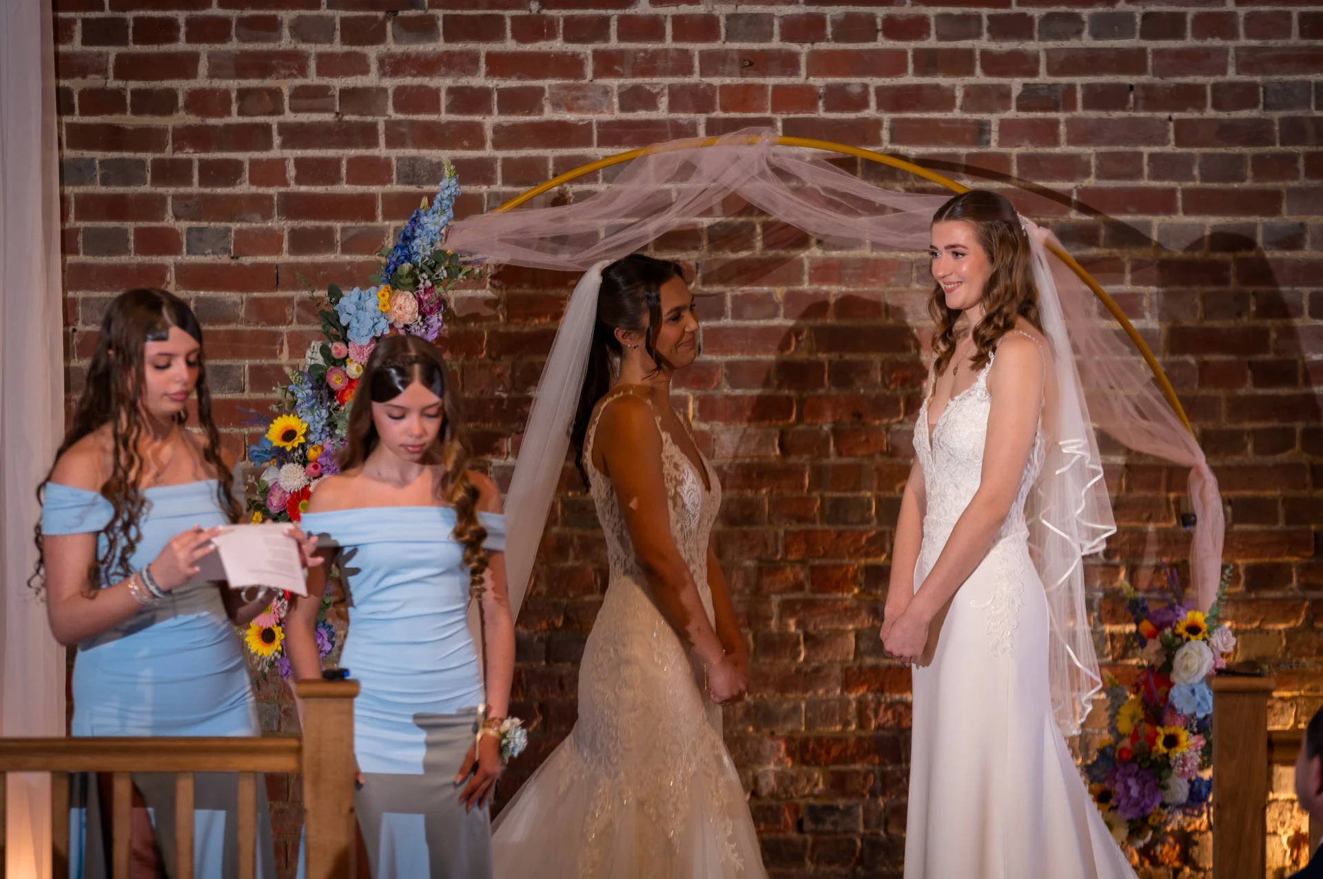 Two brides in white wedding gowns exchange loving glances during their indoor ceremony before a decorative arch and exposed brick wall, accompanied by bridesmaids in blue dresses.