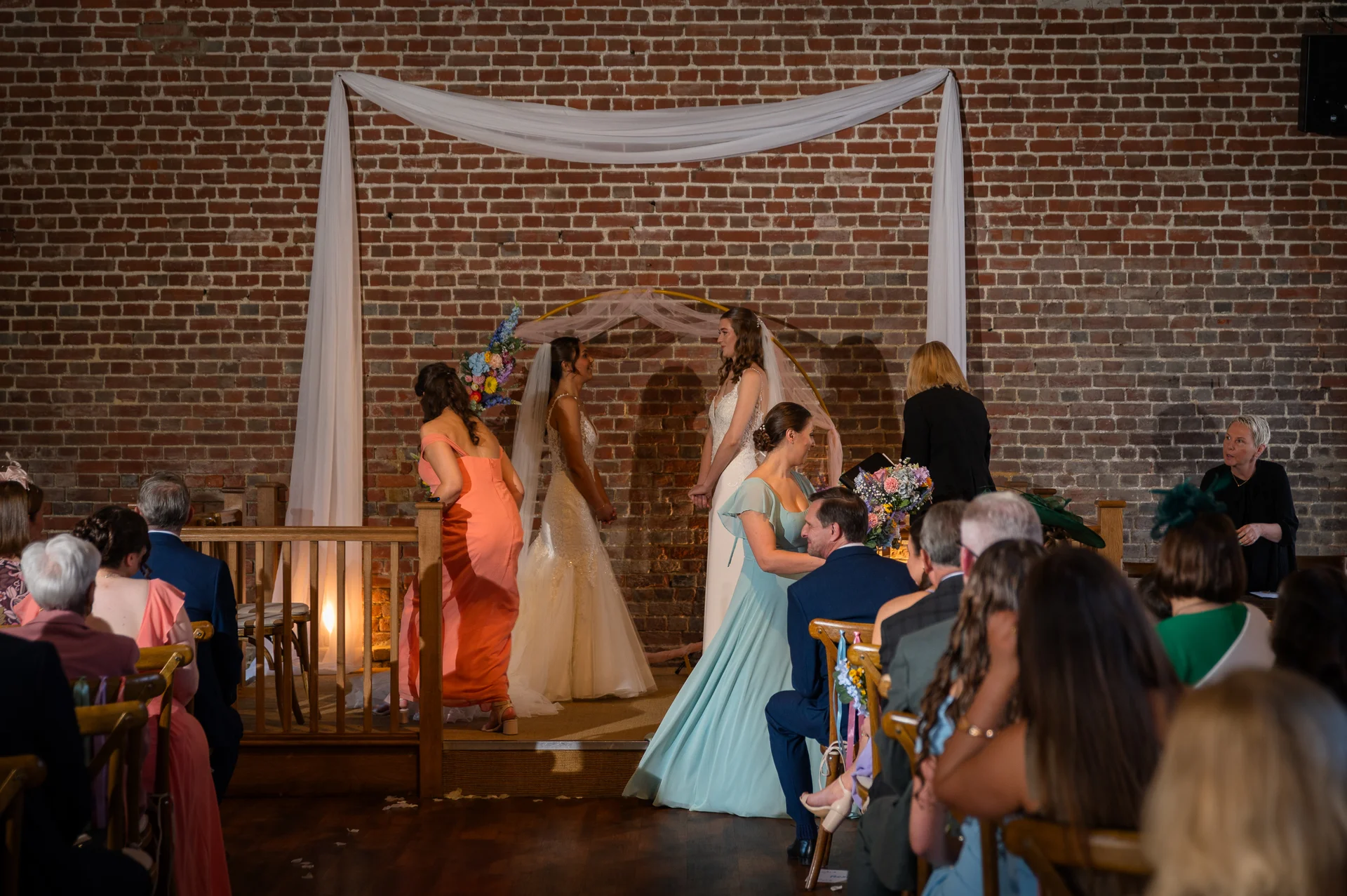 Two brides stand facing each other during their wedding ceremony in front of an exposed brick wall draped with white fabric.