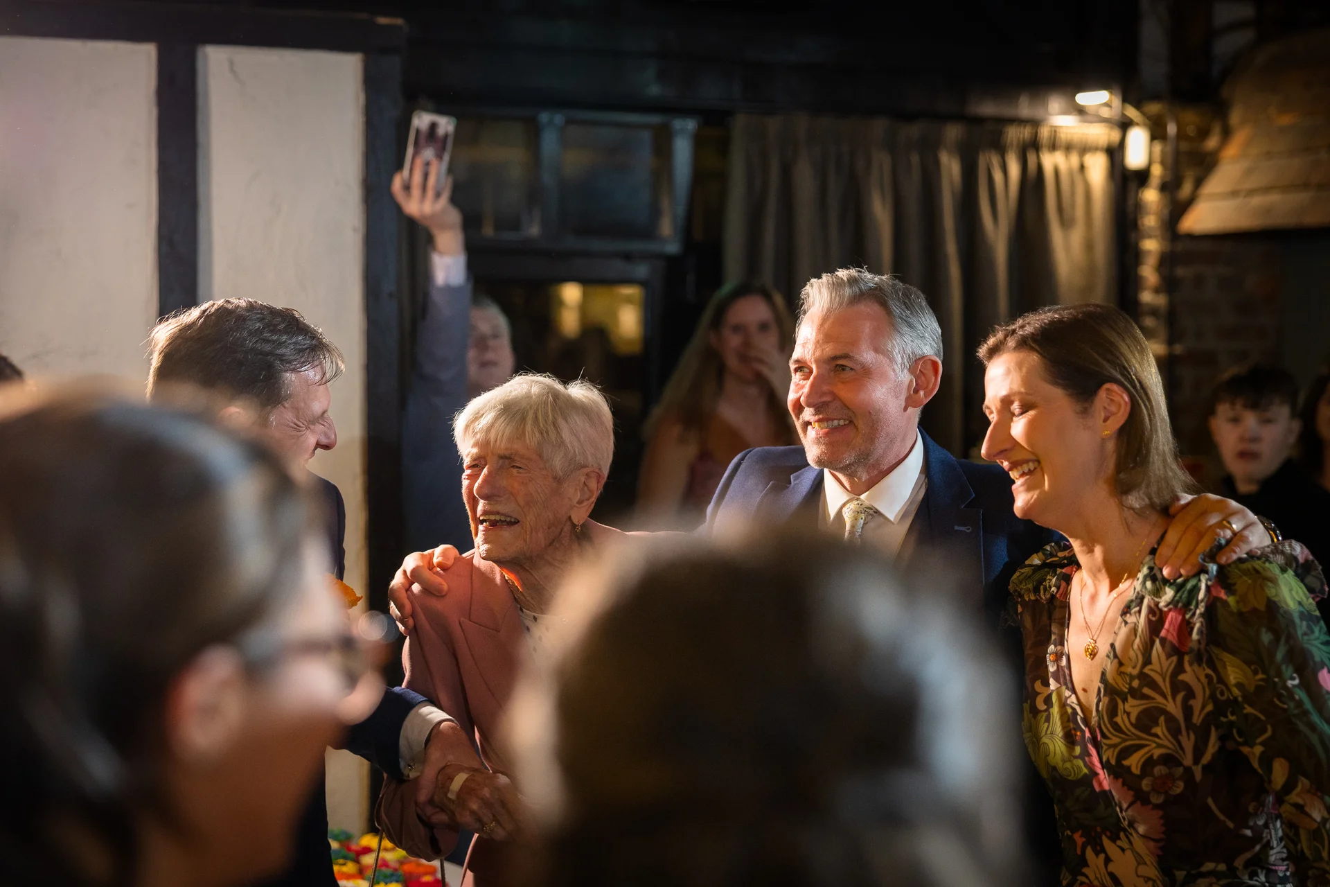 An elderly woman laughs joyfully while surrounded by smiling wedding guests and family members in a warm indoor setting.