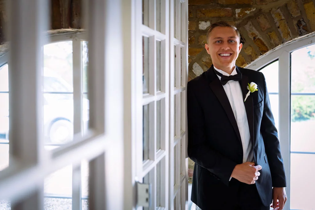 A smiling groom wearing a classic black tuxedo and a white rose boutonniere poses near a white panelled doorway with exposed brickwork.