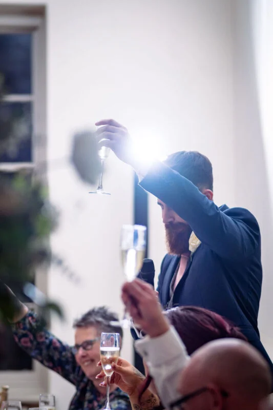 A bearded groom in a navy suit raises his glass to propose a toast during the wedding speeches, with guests raising their own glasses in celebration.