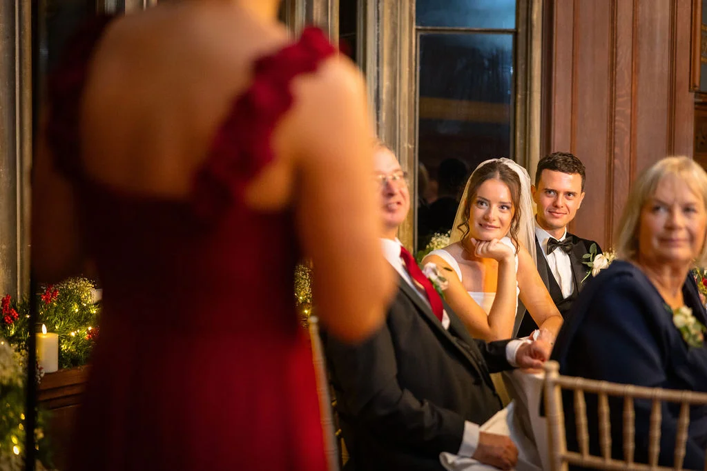 A guest in a red dress delivers a wedding speech while the bride, groom, and family members listen attentively from the top table.
