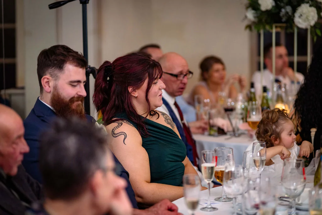 Wedding guests seated at a round dining table during the wedding breakfast, listening intently and smiling during speeches.