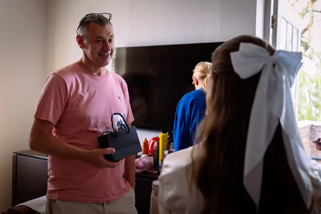 A smiling father holding a small gift bag while talking to his daughter during morning bridal preparations.