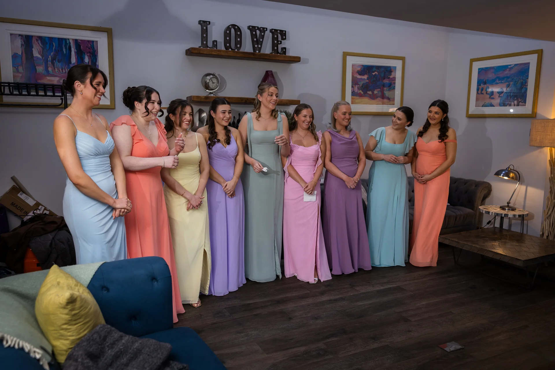 A group of nine bridesmaids in pastel-coloured mismatched dresses standing together indoors and smiling.