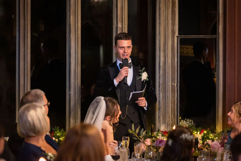A groom in a velvet tuxedo delivers his speech holding a microphone while standing before large dark windows at an evening wedding reception.