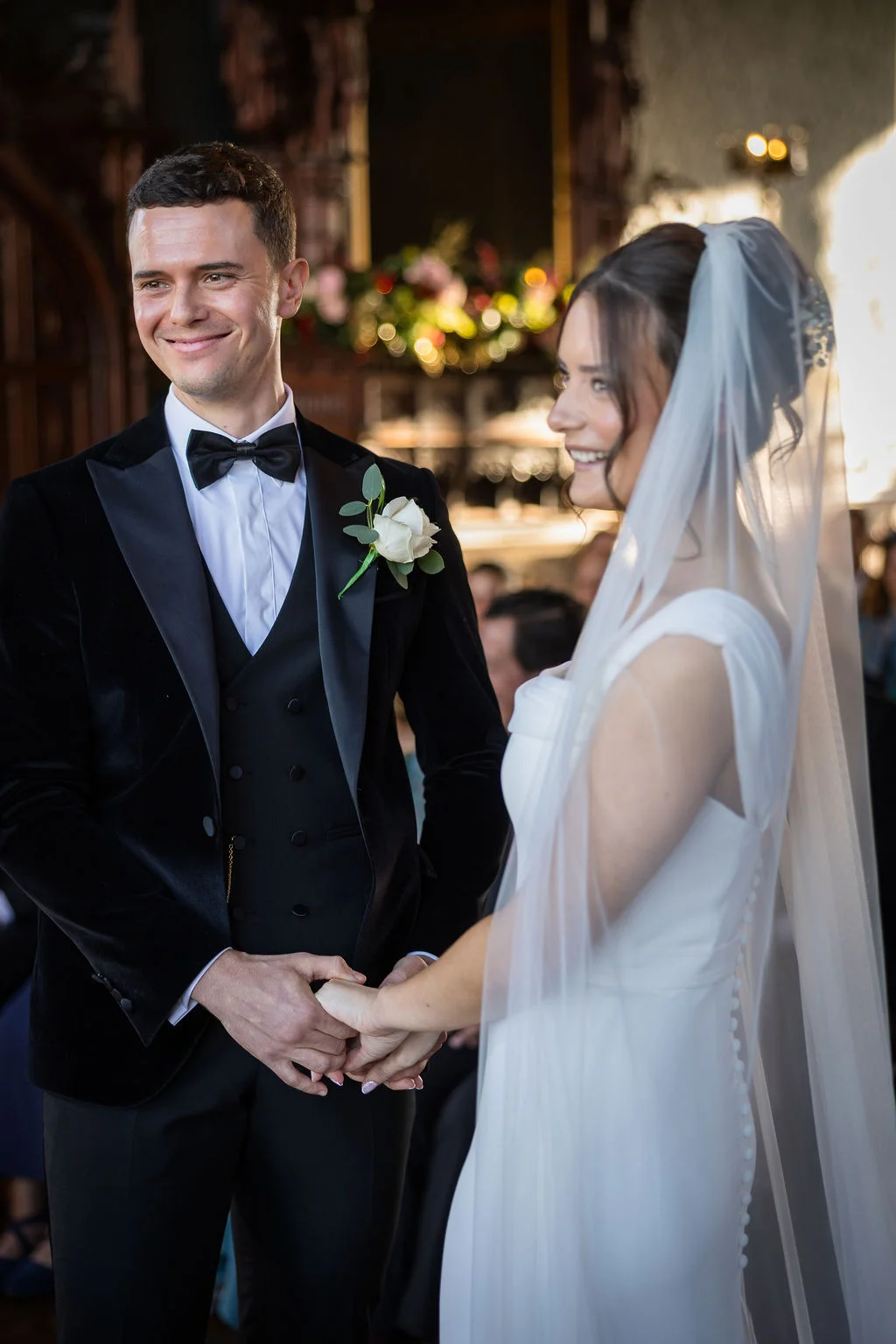 A smiling bride and groom hold hands during their indoor wedding ceremony, with the groom wearing a black velvet tuxedo and the bride in a sleek white gown and veil.