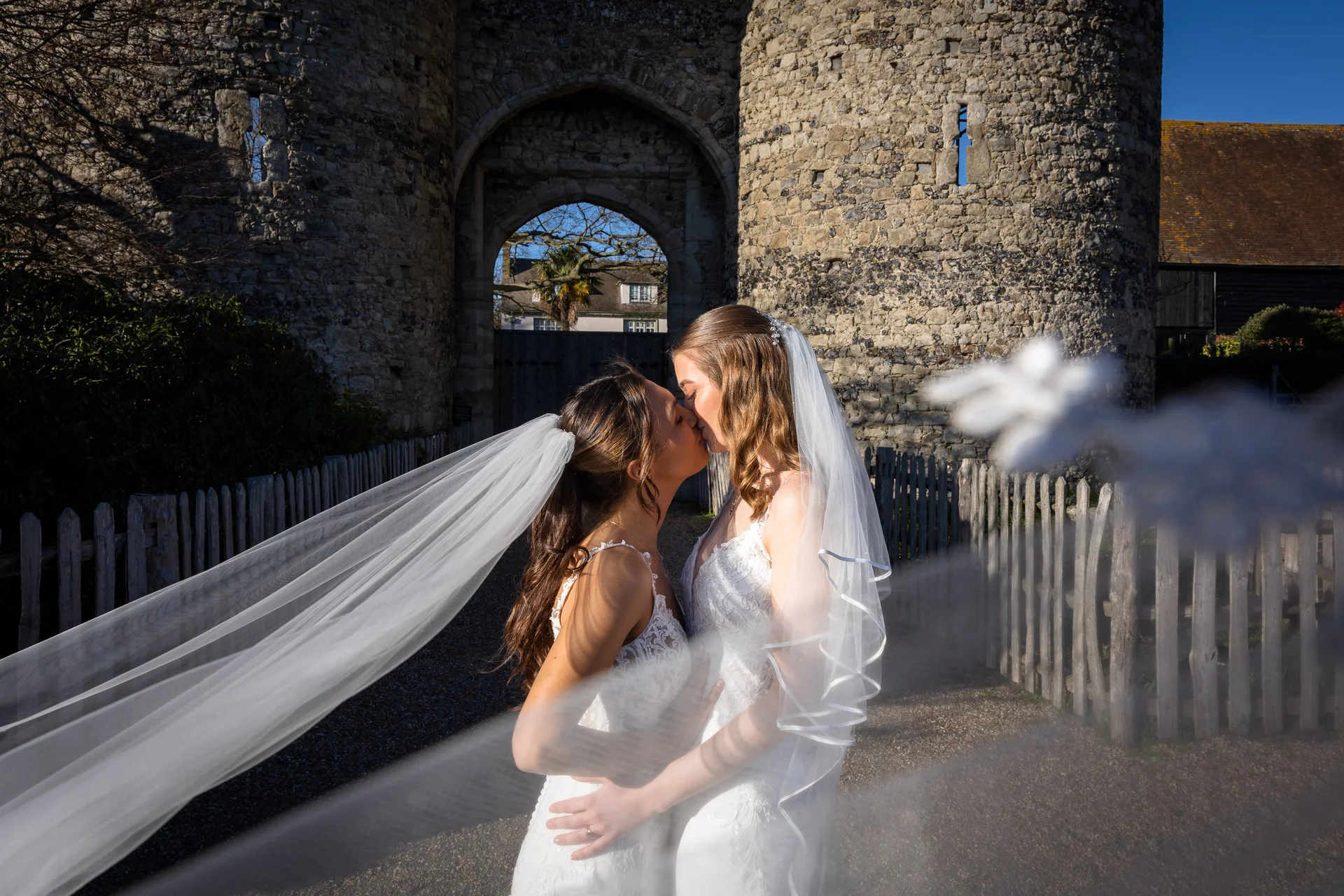 Two brides share a tender kiss in front of a historic stone castle gateway, framed dramatically by a flowing veil in the foreground.