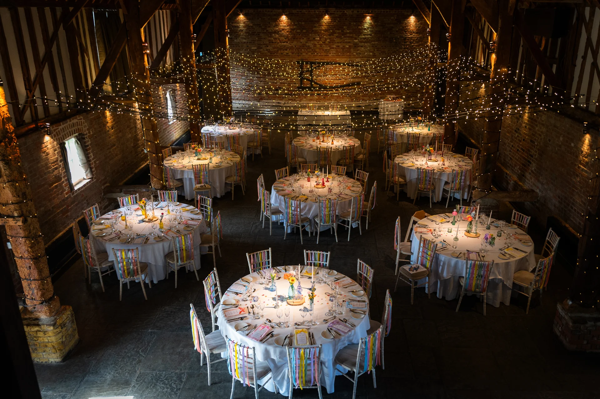 An elevated interior view of a rustic tithe barn wedding reception featuring numerous round tables decorated with floral centrepieces and colorful ribbon-tied chairs beneath an extensive canopy of twinkling fairy lights.