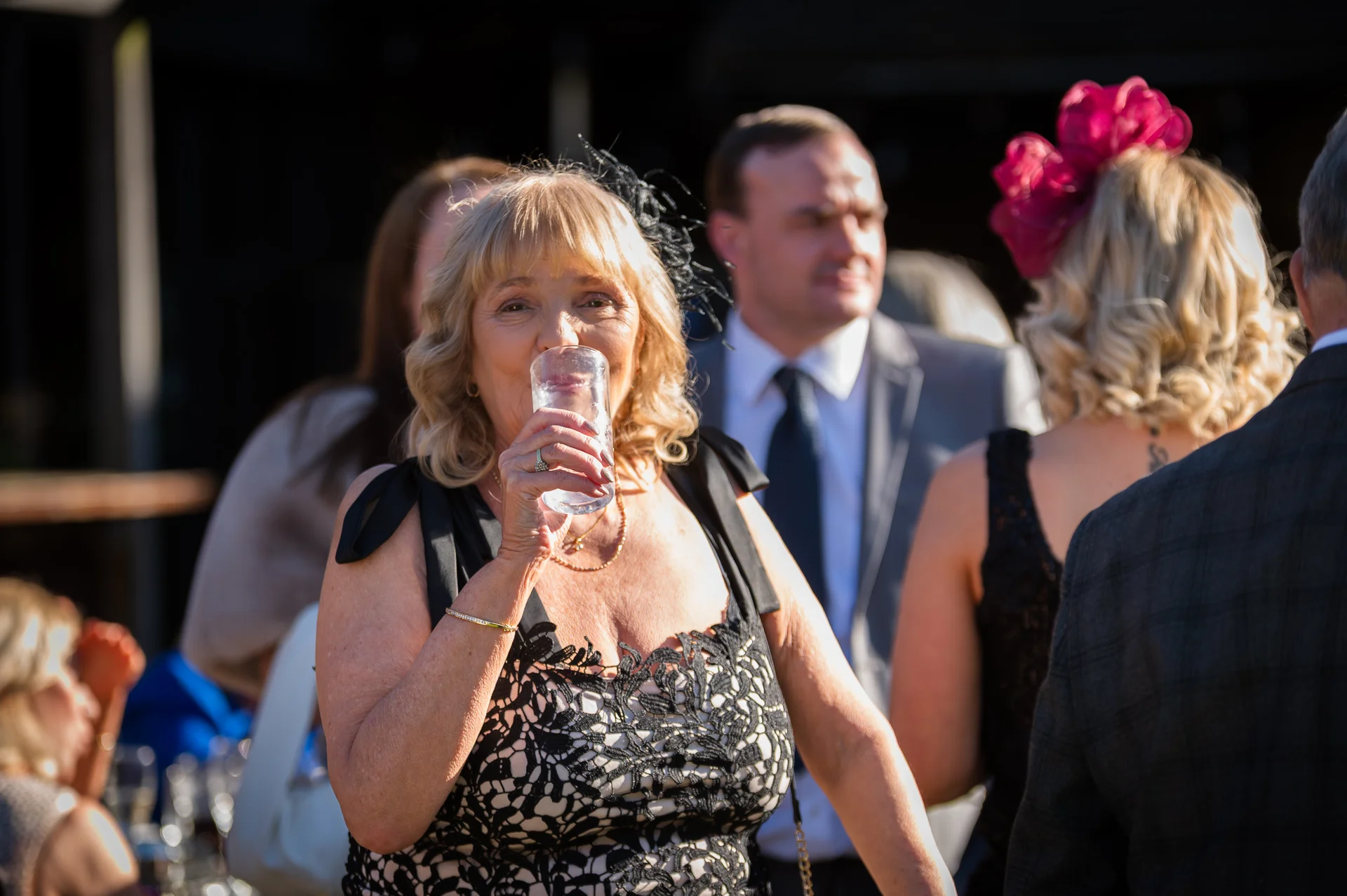 A mature wedding guest in a black lace dress drinks from a glass during an outdoor reception while other guests mingle in the background.
