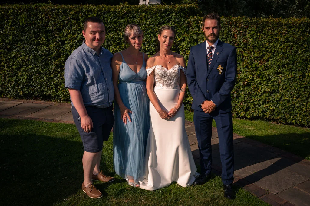 A formal group portrait of a bride, groom, and two guests posing outdoors in front of a neatly trimmed garden hedge on a sunny afternoon.