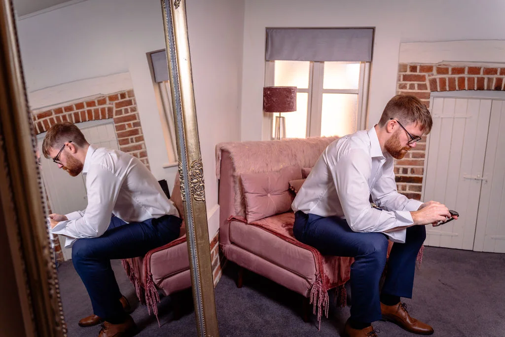 A groom sits on a pink velvet sofa in front of a mirror, adjusting his cufflinks while getting ready in a room with exposed brick walls.