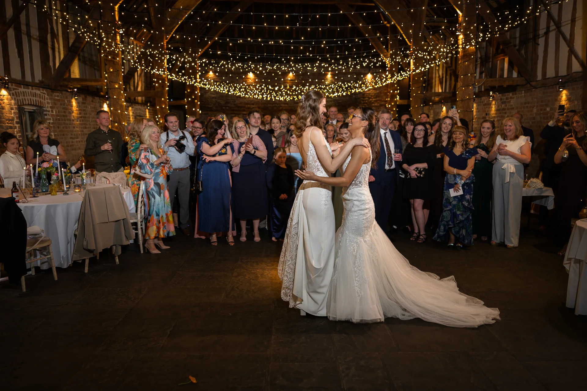 Two brides share their first dance in a rustic oak-framed barn adorned with a magnificent canopy of twinkling fairy lights while guests watch on happily.