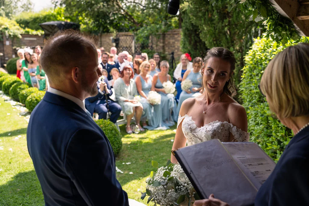 An outdoor wedding ceremony takes place in a sunny garden, with the bride and groom standing before the celebrant as guests look on.
