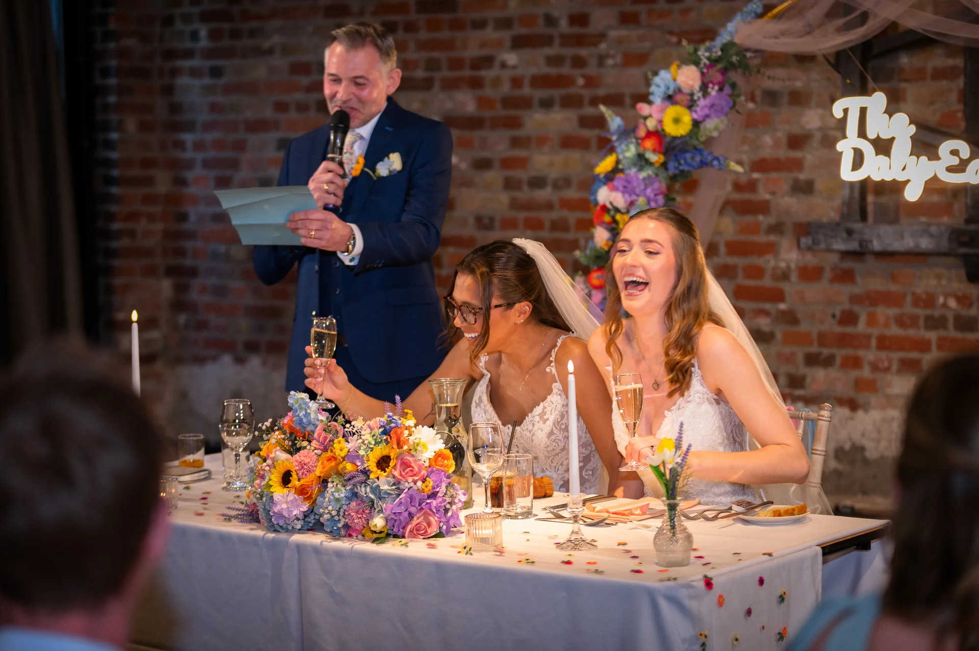 Two smiling brides sit at a beautifully decorated top table sharing a joyous moment while a family member delivers a heartfelt speech during the wedding reception.