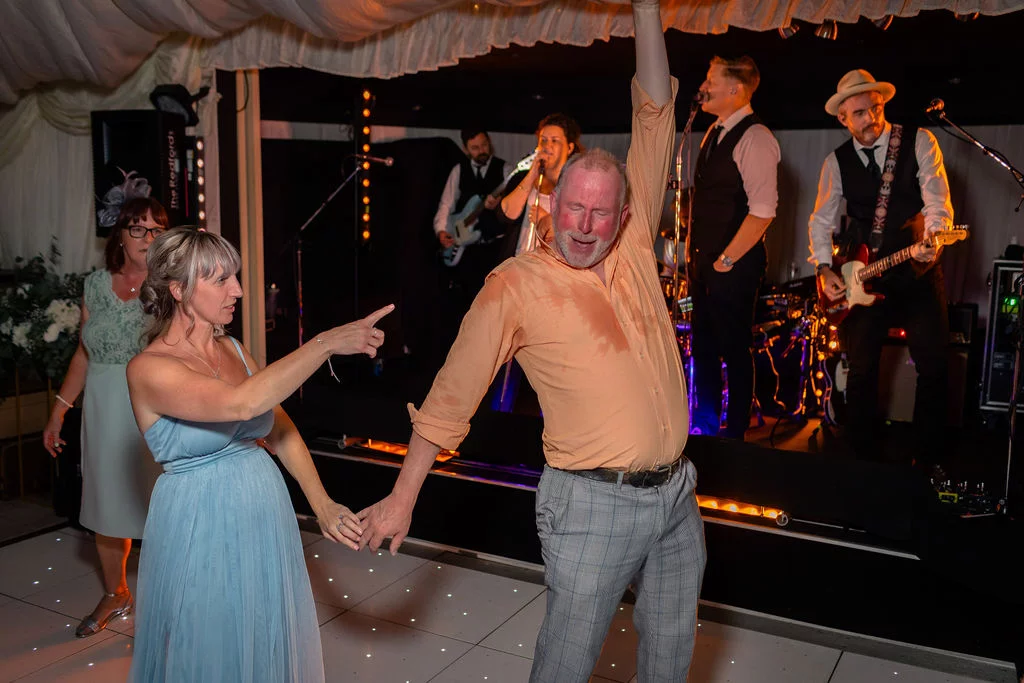 A guest in a peach shirt and grey checked trousers dances enthusiastically on a lighted dance floor while a live band performs on stage behind him.