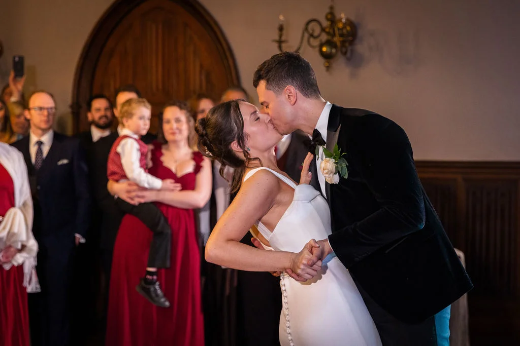 Newlyweds share a romantic dip and kiss during their first dance while wedding guests watch closely in a historic venue.