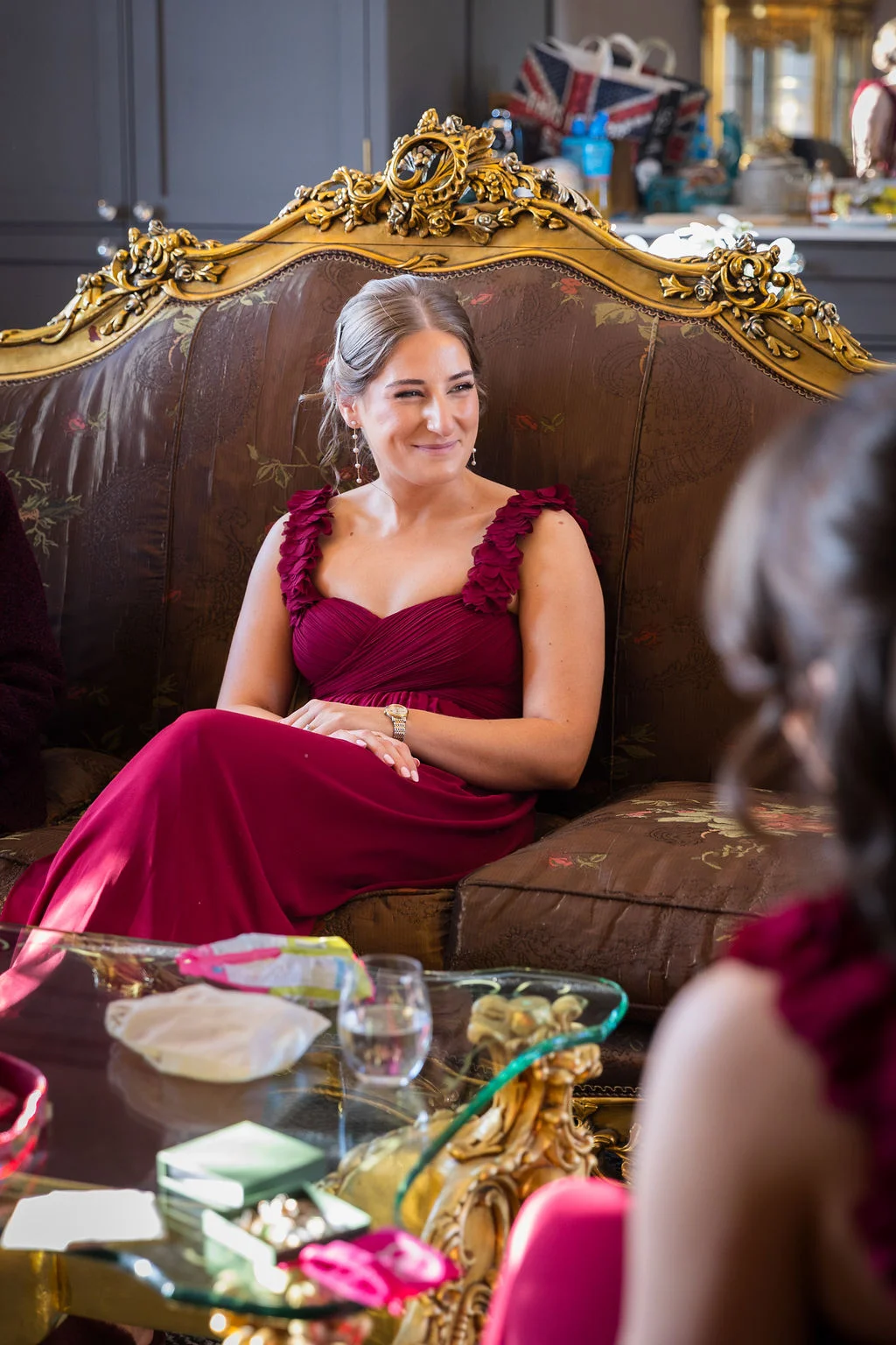 A bridesmaid in a burgundy dress sits smiling on an ornate vintage sofa while getting ready.