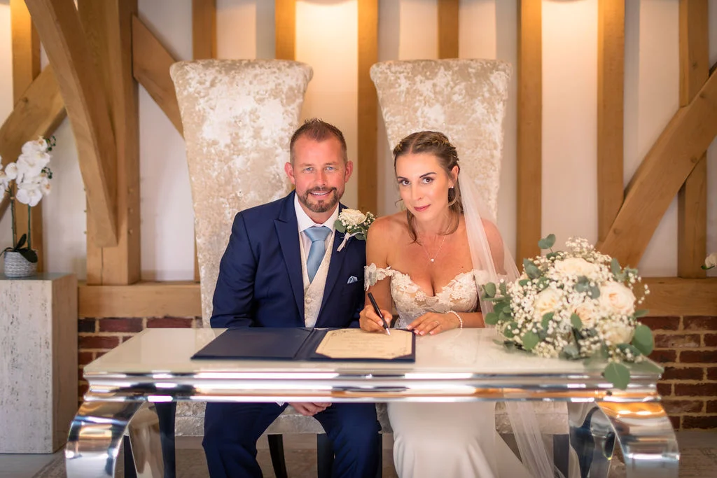 The bride and groom sit together at a sleek white table in an oak-framed barn to sign their wedding register.