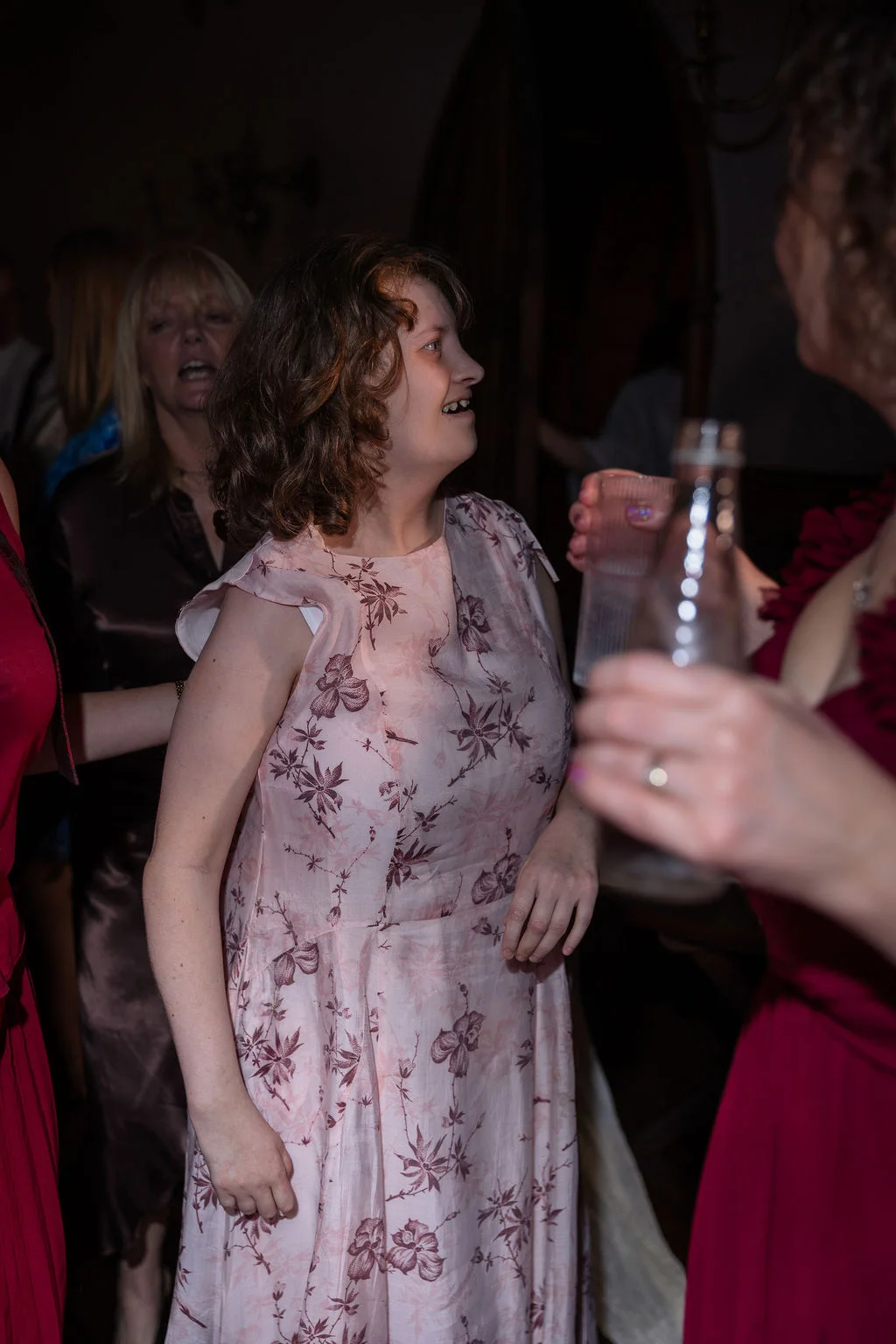 A guest in a floral pink dress smiles and enjoys the evening wedding reception surrounded by mingling family and friends.