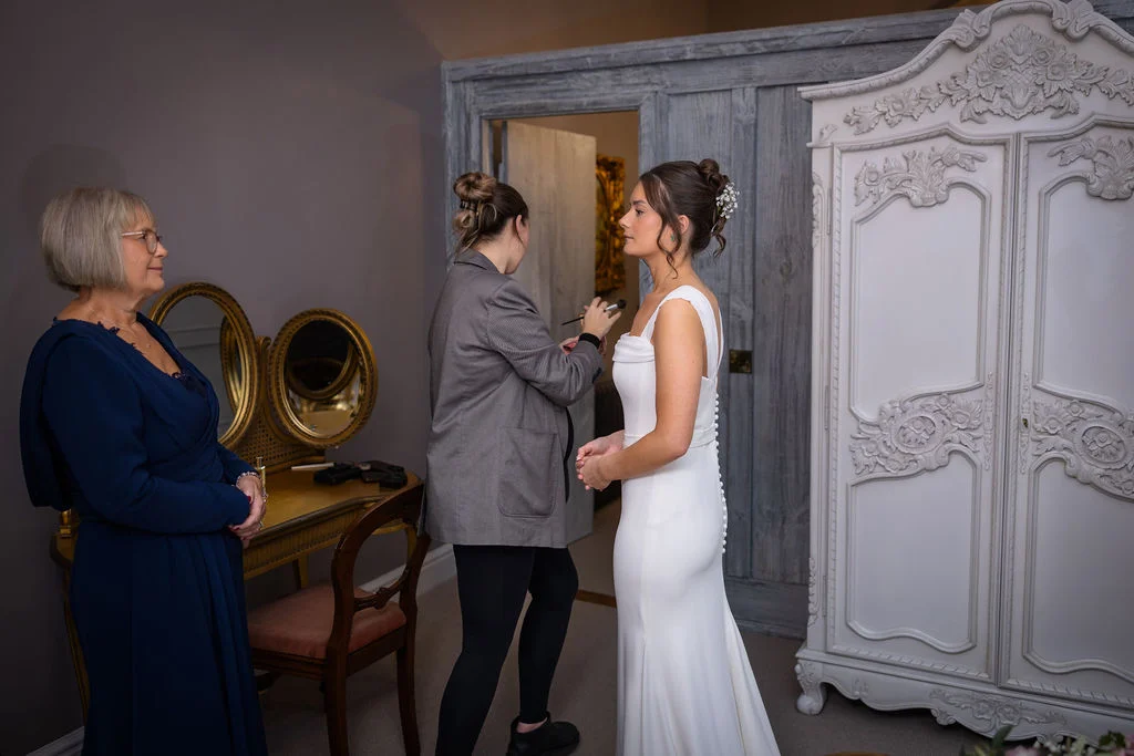 A bride in a sleek white crepe column gown stands getting her final makeup touches applied by a makeup artist, while her mother looks on admiringly in a room with a vintage wardrobe and vanity.
