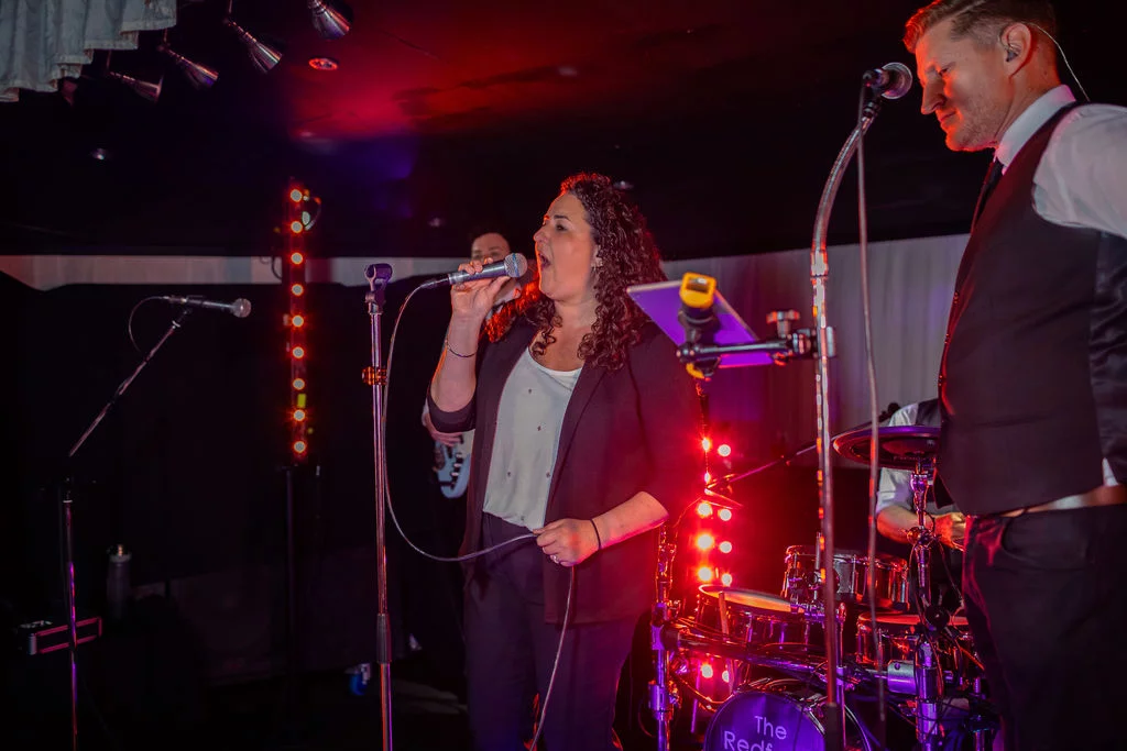 A live band performs on stage with vibrant stage lighting, featuring a female lead singer and a male singer and percussionist.