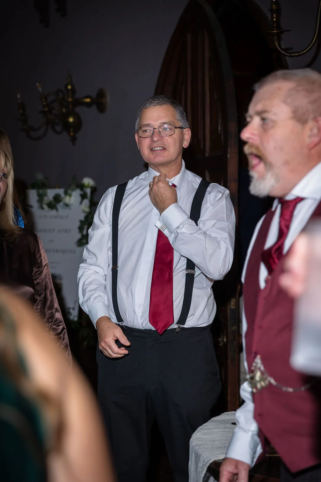A wedding guest in a white shirt, red tie, and black trousers with braces adjusts his collar during an indoor reception.