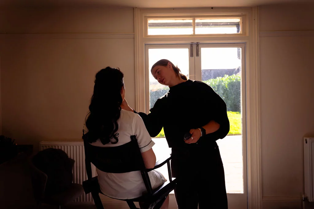 A makeup artist delicately applies makeup to a bride seated in a chair, beautifully silhouetted against bright window light during morning preparations.