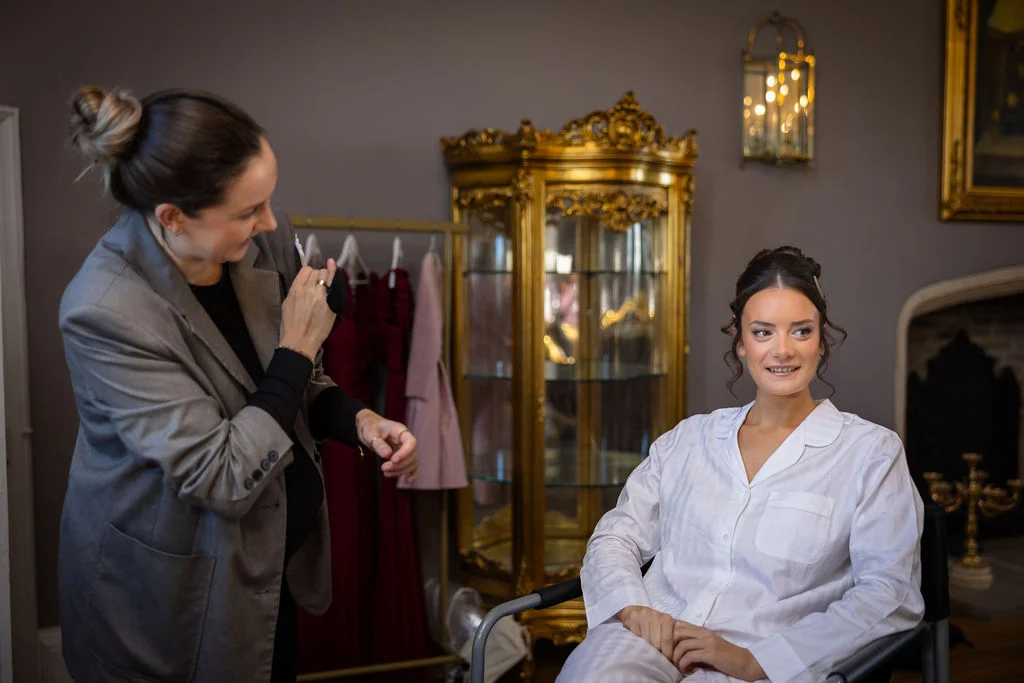 A bride wearing matching pyjamas sits smiling as a makeup artist puts the finishing touches to her hair during morning bridal preparations.