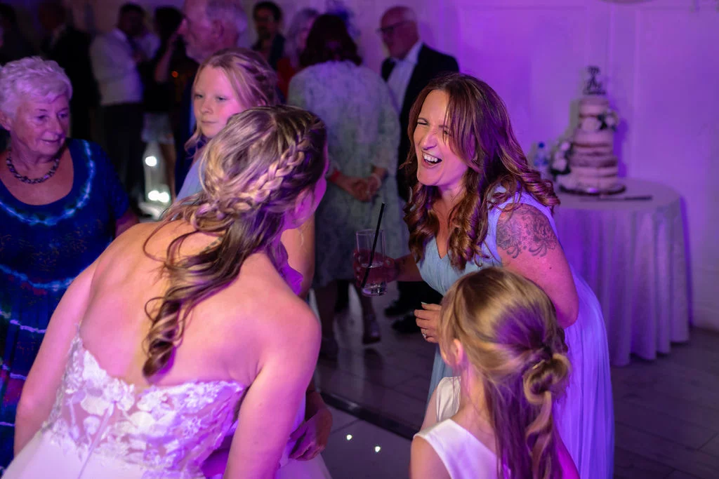 A bride in a lace strapless gown with a braided updo laughs joyfully with wedding guests under vibrant purple reception lighting.