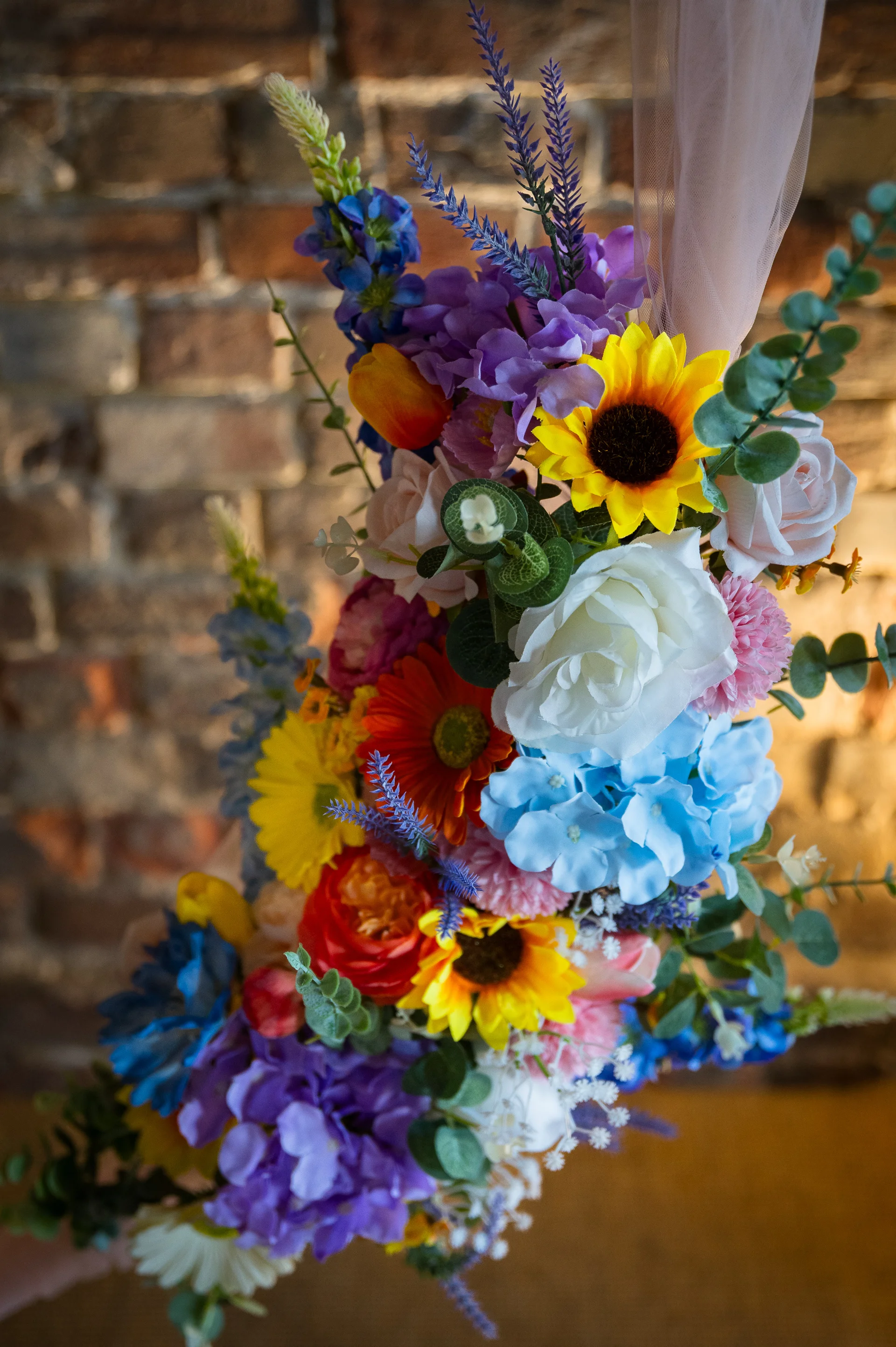 A vibrant floral arrangement featuring colourful sunflowers, blue hydrangeas, white roses, and lavender against an exposed brick wall background.