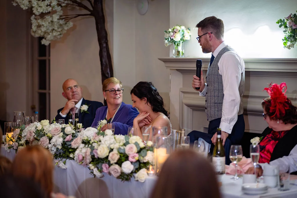A groom delivers a wedding speech standing near the top table while the bride and family members listen and smile.
