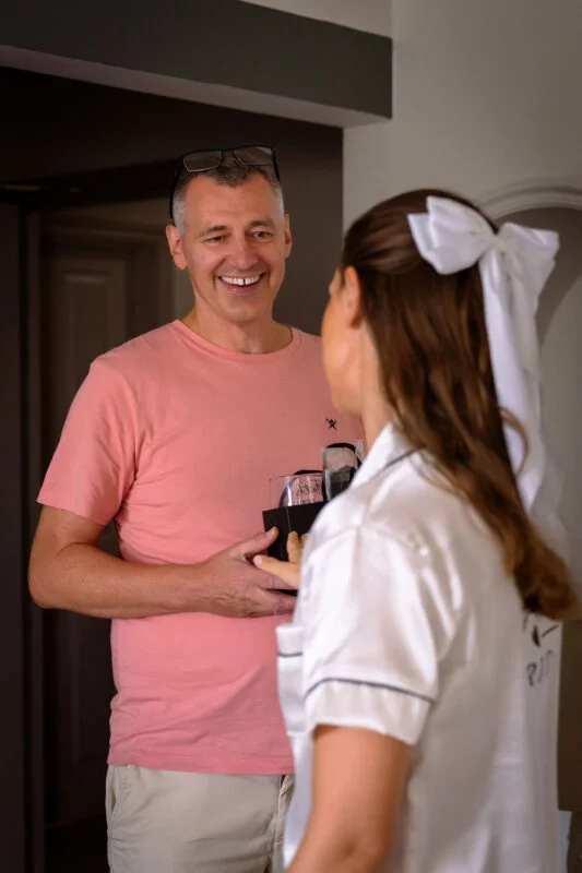 A smiling father wearing a pink t-shirt holds a gift while interacting with a bride in white pyjamas and a large hair bow during morning preparations.