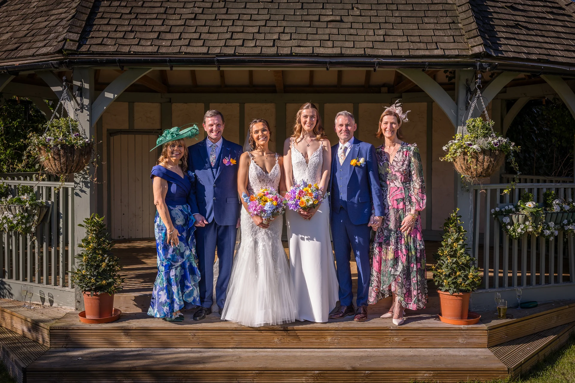 A smiling family group and a same sex couple pose together on the wooden steps of a gazebo, holding colourful bouquets and dressed in wedding attire.