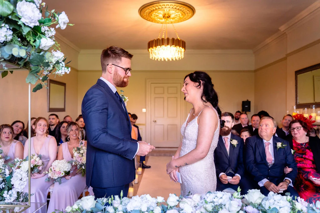 A groom in a navy suit and a bride in a sparkling strap gown exchange vows during an indoor civil ceremony surrounded by seated guests and floral arrangements.