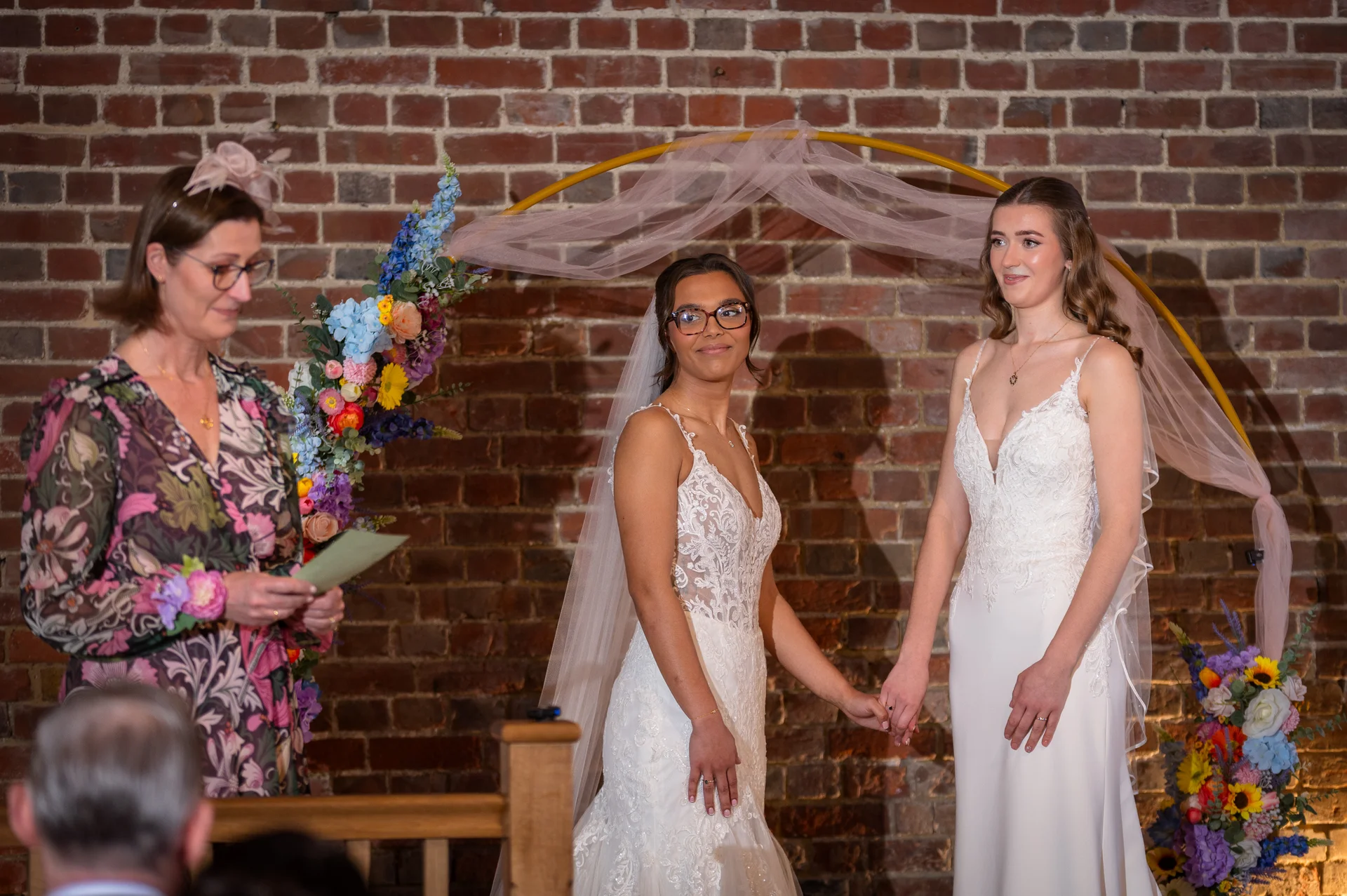 Two smiling brides stand hand in hand at the altar during an indoor ceremony, framed by a floral arch and an exposed brick wall.