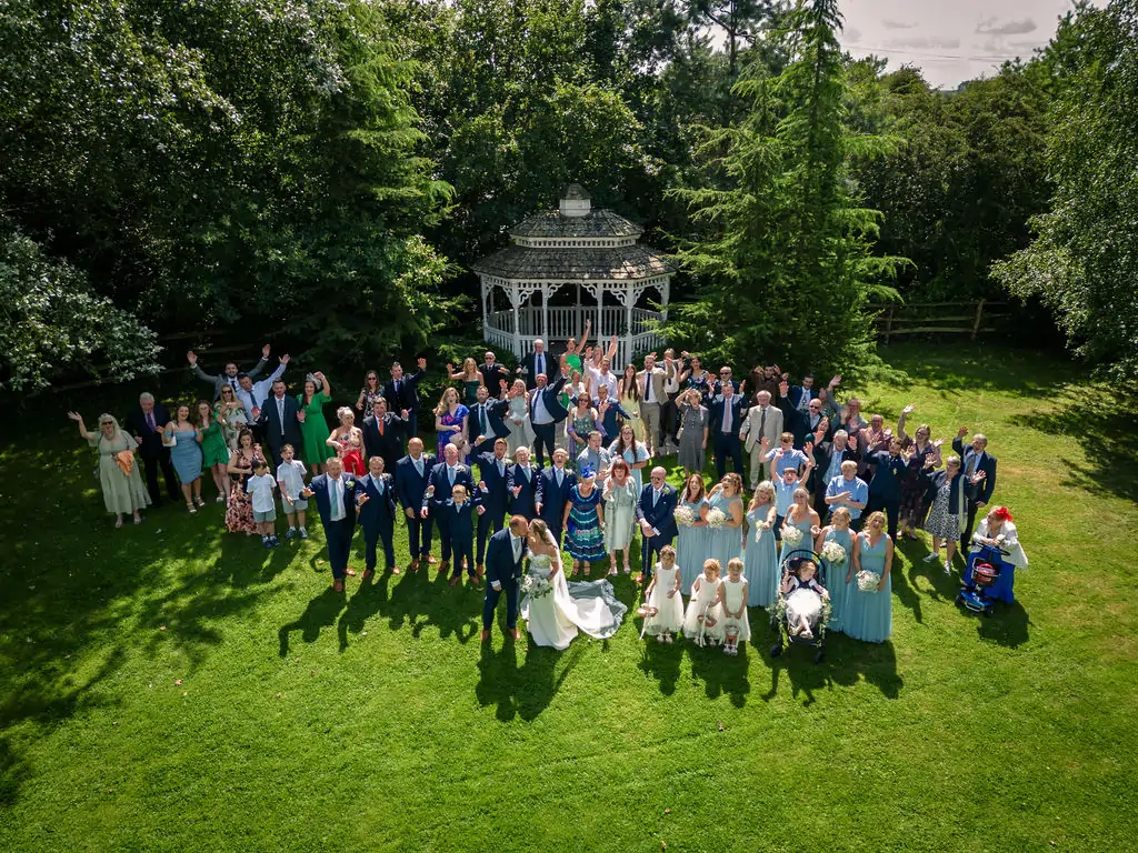 An aerial drone shot captures a large wedding party and guests cheering and waving on a lush green lawn in front of a white gazebo.