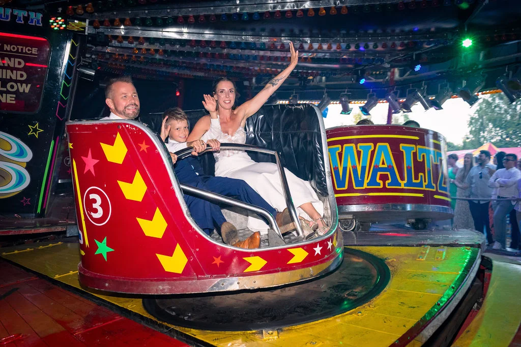 A joyful bride and groom laughing while riding a waltzer fairground attraction with a young boy during their festival-themed wedding reception.