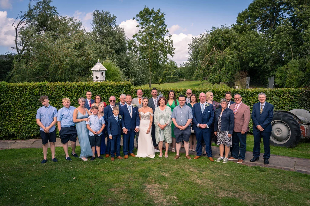 A large wedding party and family group pose for a formal portrait outdoors on a manicured lawn beside a green hedge and a vintage tractor.