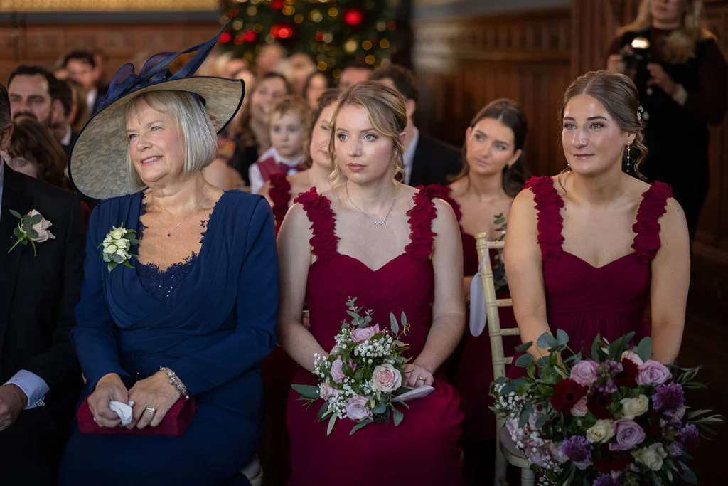 The mother of the bride in a blue dress and large hat sits alongside bridesmaids wearing matching burgundy dresses and holding bouquets during the wedding ceremony.