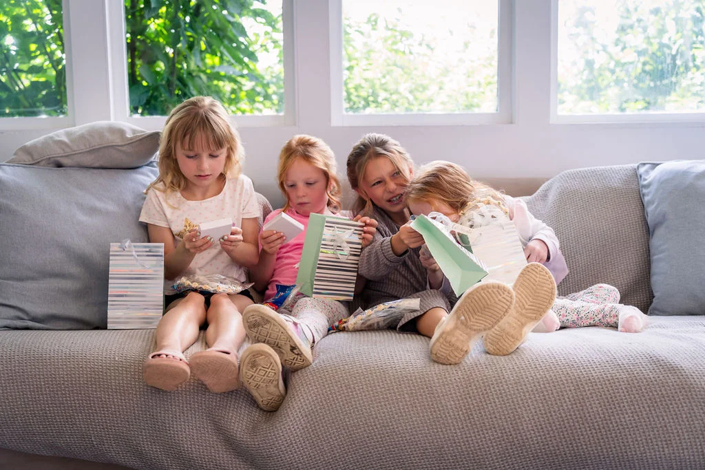 Four young flower girls sit together on a neutral sofa indoors, excitedly opening small gift bags and boxes near a bright window.