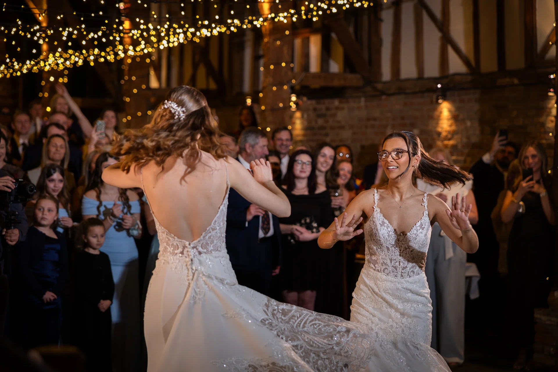 Two smiling brides in detailed lace wedding dresses joyfully twirl on the dance floor of a rustic barn venue illuminated by fairy lights.