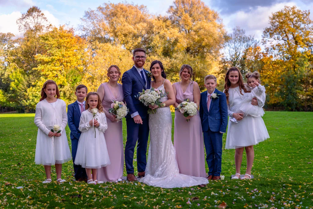 A bridal party including bridesmaids in pink dresses, groomsmen, and flower girls poses with the newly married couple on a grassy lawn surrounded by autumnal trees.