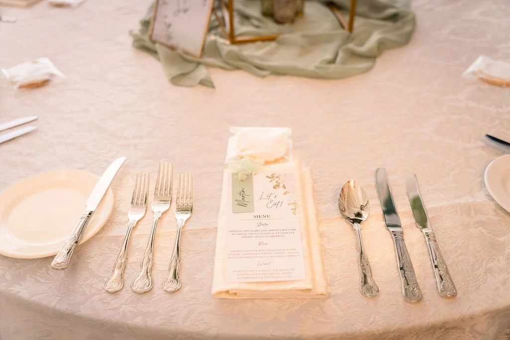 An elegantly styled wedding place setting featuring a printed menu on a folded napkin, shiny silverware, and a delicate sage green table runner on a round table.