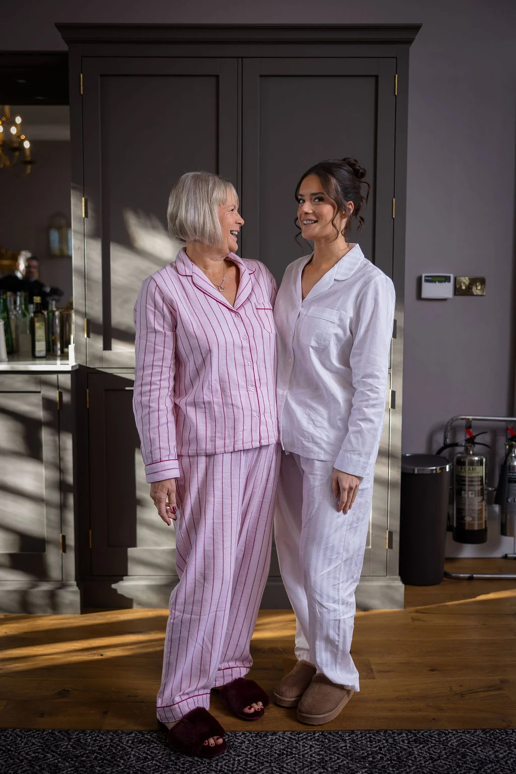 A smiling bride and her mother wearing matching pyjamas share a joyful moment together indoors during morning bridal preparations.