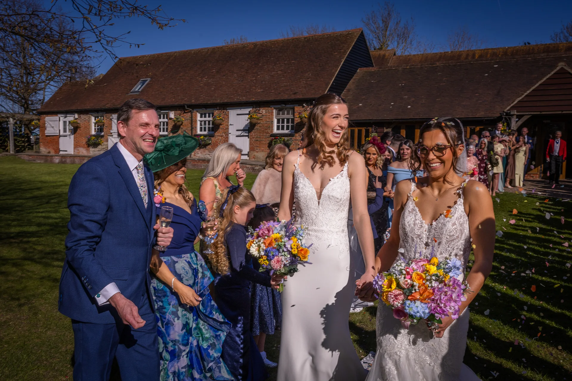 Two smiling brides holding colorful wildflower bouquets walk hand in hand through a joyful confetti exit outside a rustic stone barn on a sunny day.