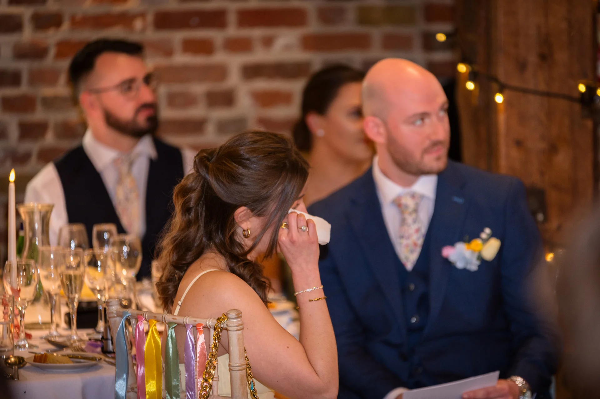 A wedding guest wiping away a tear during speeches at a reception table decorated with candles and glassware against an exposed brick wall.