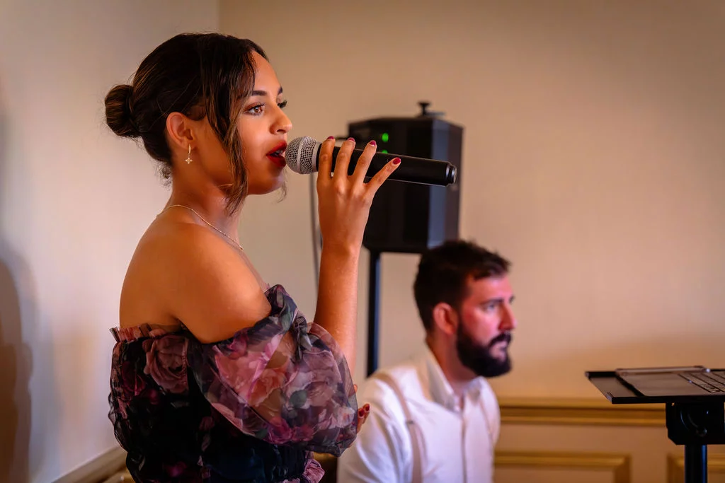 A smiling bride wearing an off-the-shoulder floral print dress sings into a microphone during the wedding reception.