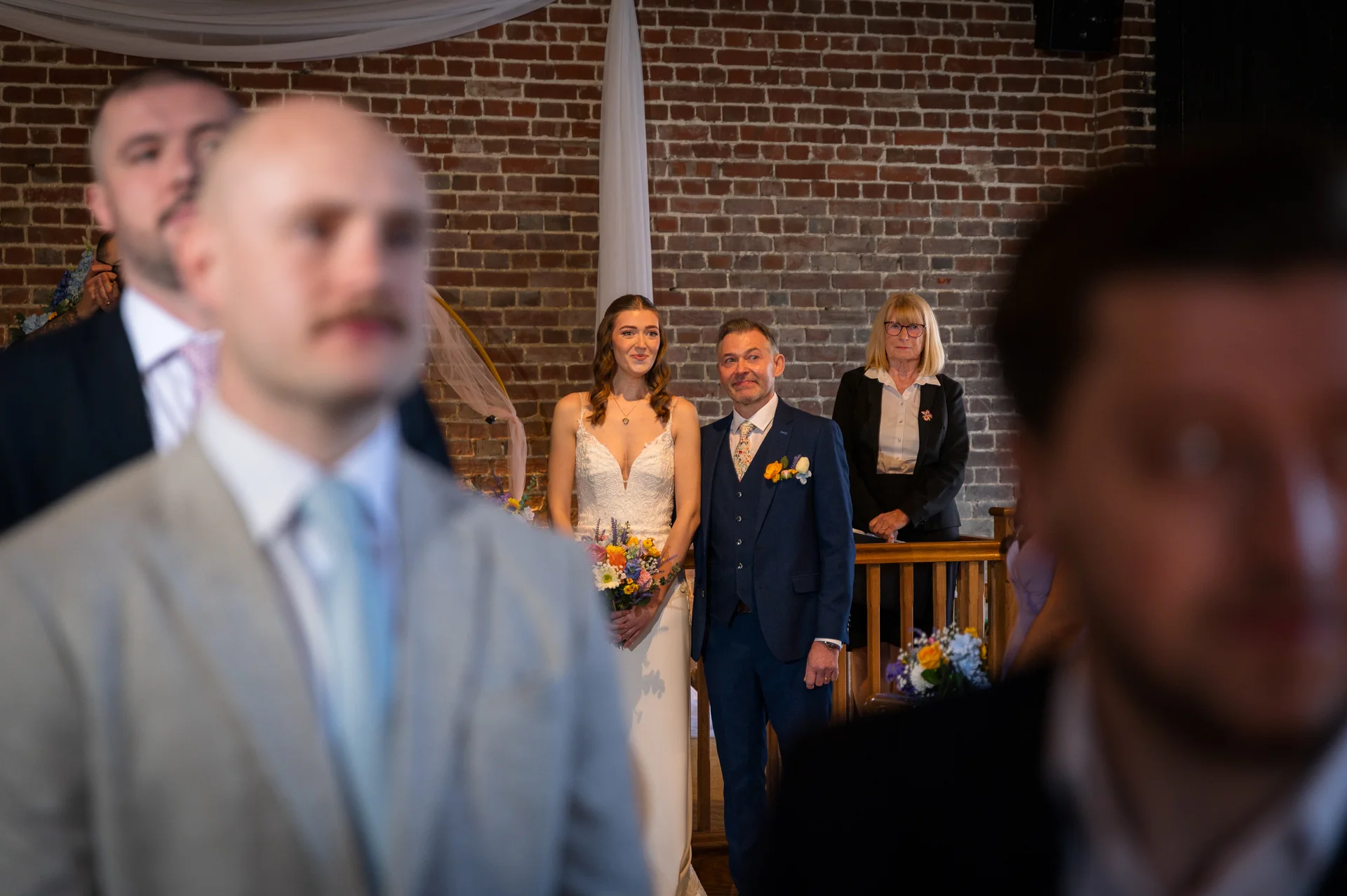 A bride in a white gown holding a bouquet stands at the altar next to a groom in a blue suit during a wedding ceremony, framed by guests in the foreground.