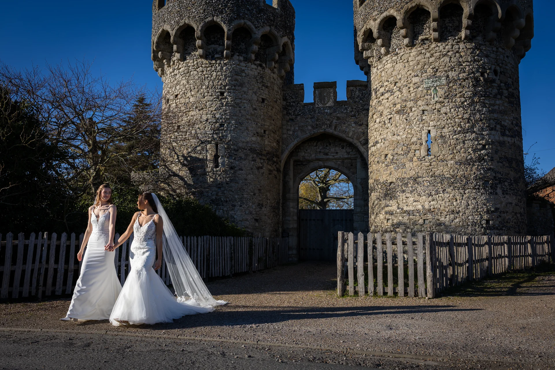 Two brides in white wedding dresses walk hand in hand along a gravel path in front of a historic stone castle gatehouse under a clear blue sky.